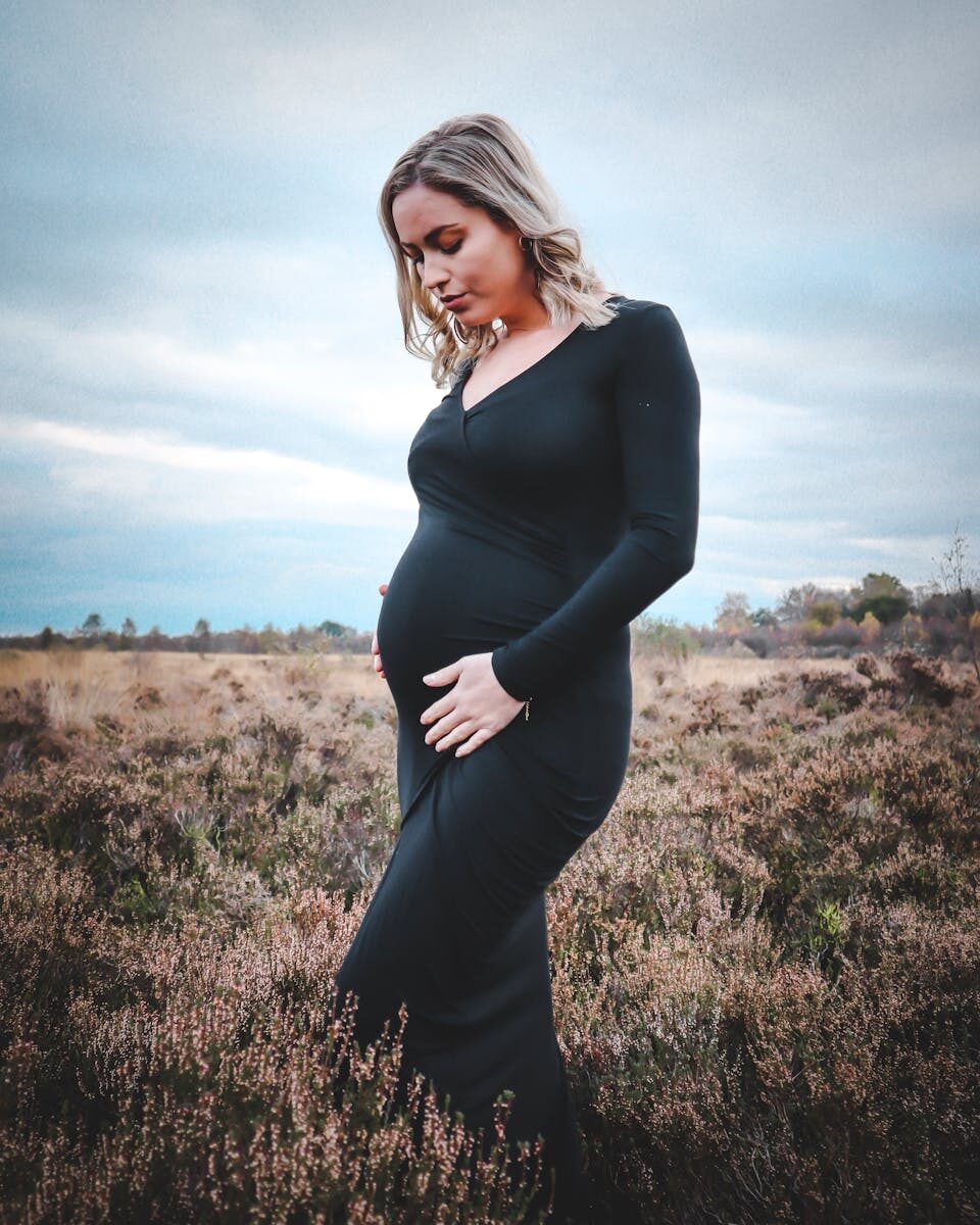 Pregnant woman in black dress posing outdoors in a tranquil field setting. This demonstrates our birth reflection service. 